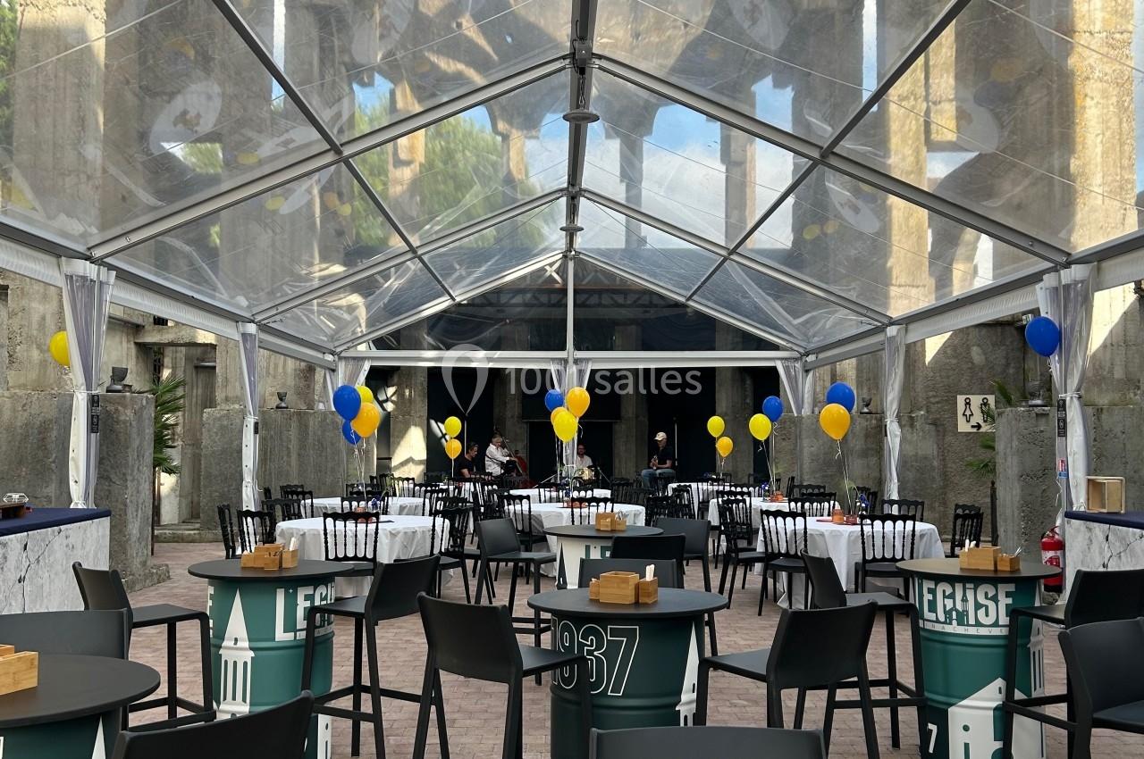Salle extérieure sous une tente transparente avec tables, chaises et décorations aux couleurs bleu et jaune.