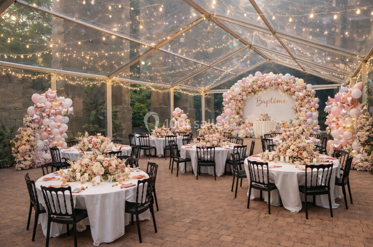 Salle de réception décorée pour un baptême, avec tables rondes, nappes blanches, fleurs et guirlandes lumineuses sous une…