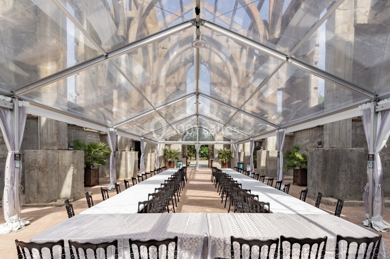 Salle extérieure sous une tente transparente, aménagée avec de longues tables blanches et des chaises noires.