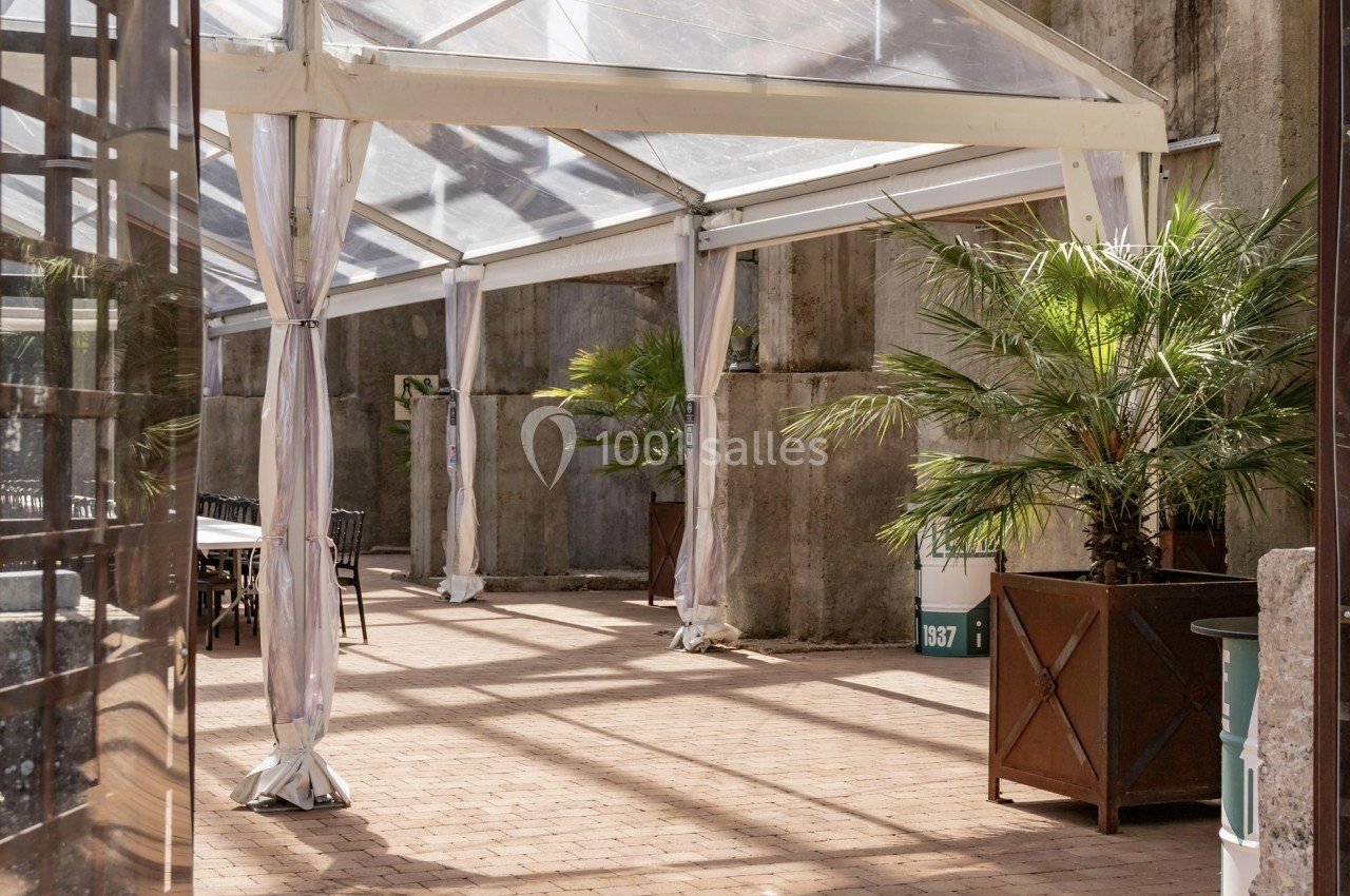 Terrasse extérieure avec sol en pavés, palmiers en pots et structure en verre et métal laissant passer la lumière.
