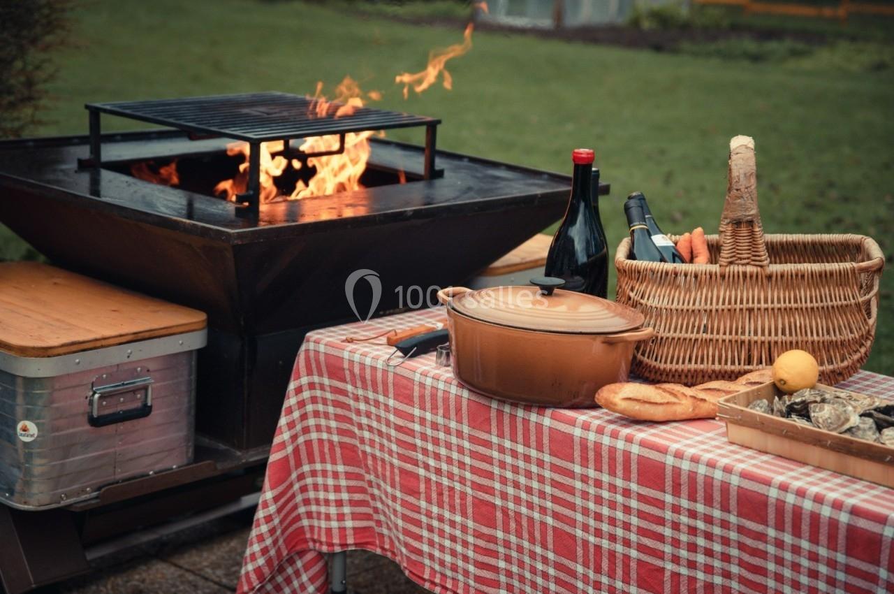 Table de pique-nique avec nappe à carreaux, panier en osier, plats, bouteilles de vin et barbecue allumé en arrière-plan.