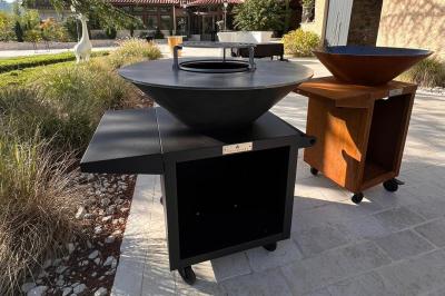 Deux personnes cuisinent des légumes colorés sur une grande plancha en extérieur, par une journée ensoleillée.