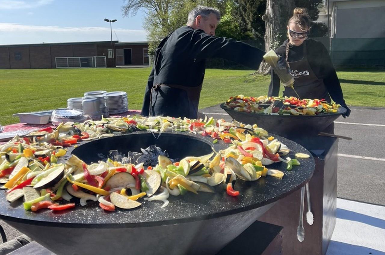 Deux personnes cuisinent des légumes colorés sur une grande plancha en extérieur, par une journée ensoleillée.