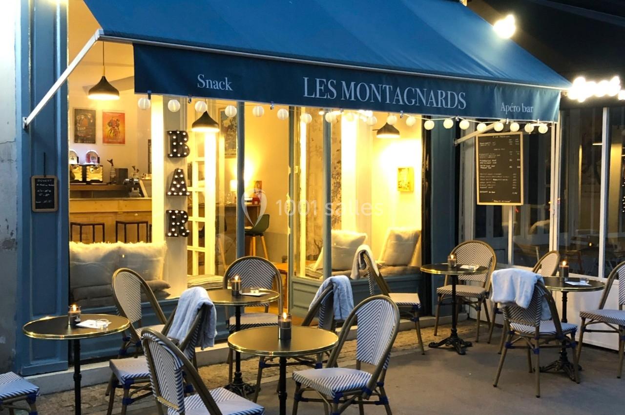 Terrasse d'un café avec chaises en osier et tables rondes, devant une façade éclairée portant l'enseigne ’Les Montagnards’.