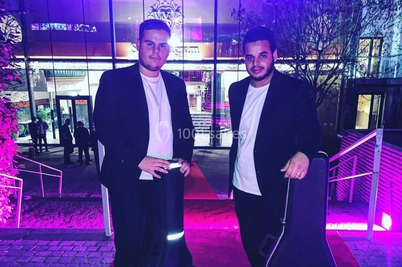 Deux hommes en tenue décontractée posent devant un bâtiment illuminé de lumières violettes.