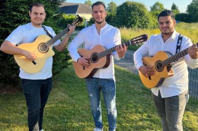 Trois hommes jouent de la guitare acoustique dans un jardin, vêtus de chemises blanches et entourés de verdure.