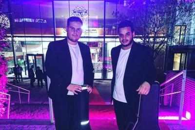 Deux hommes en costume posent devant un bâtiment éclairé par des lumières violettes, avec une guitare visible à droite.