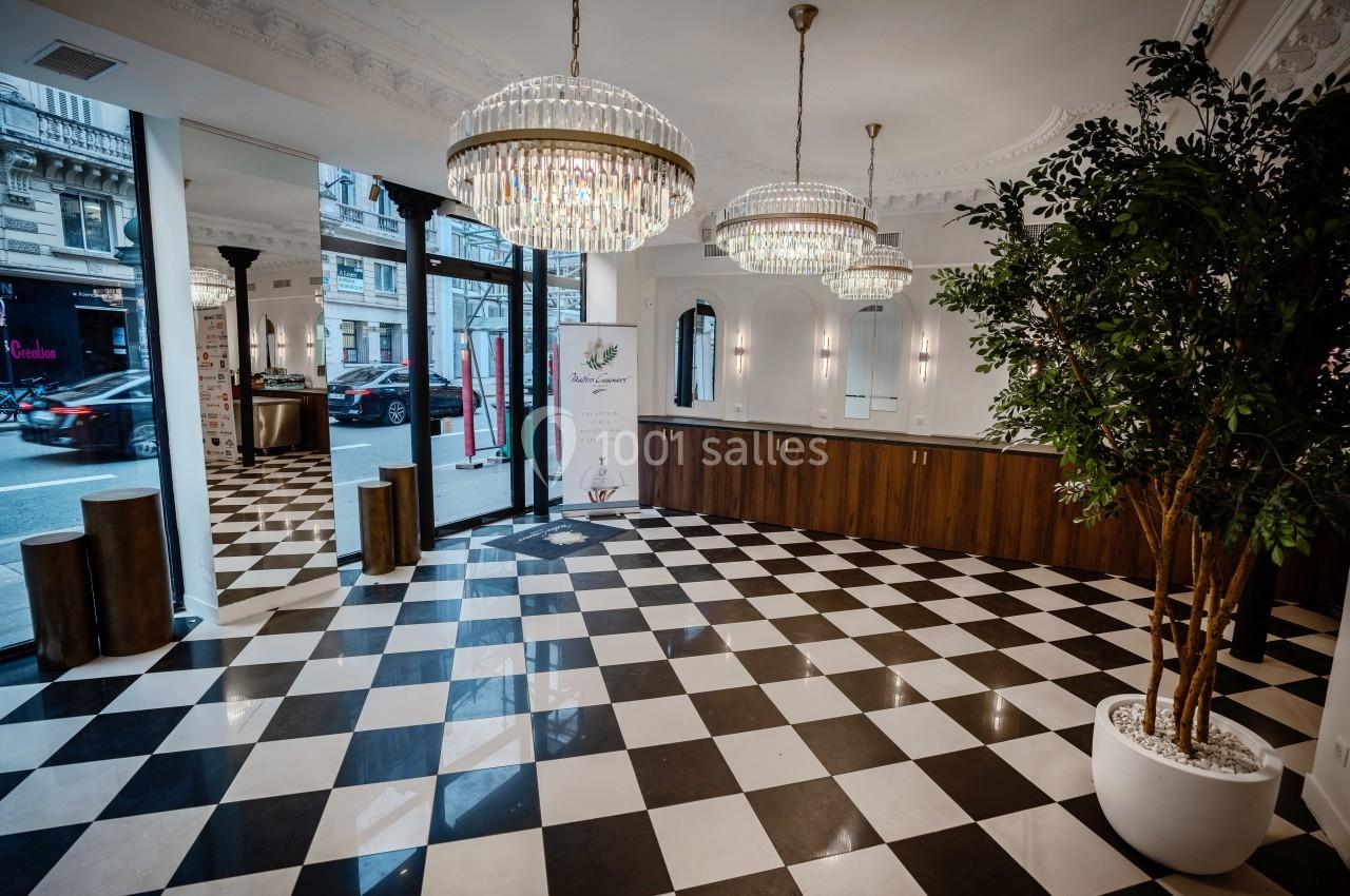 Intérieur d'un espace lumineux avec sol en damier, comptoir en bois, lustres et grandes baies vitrées donnant sur la rue.