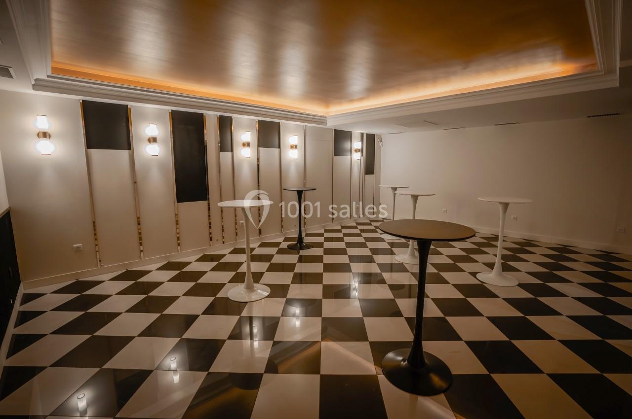 Salle vide avec sol en damier noir et blanc, tables hautes dispersées et éclairage mural moderne.