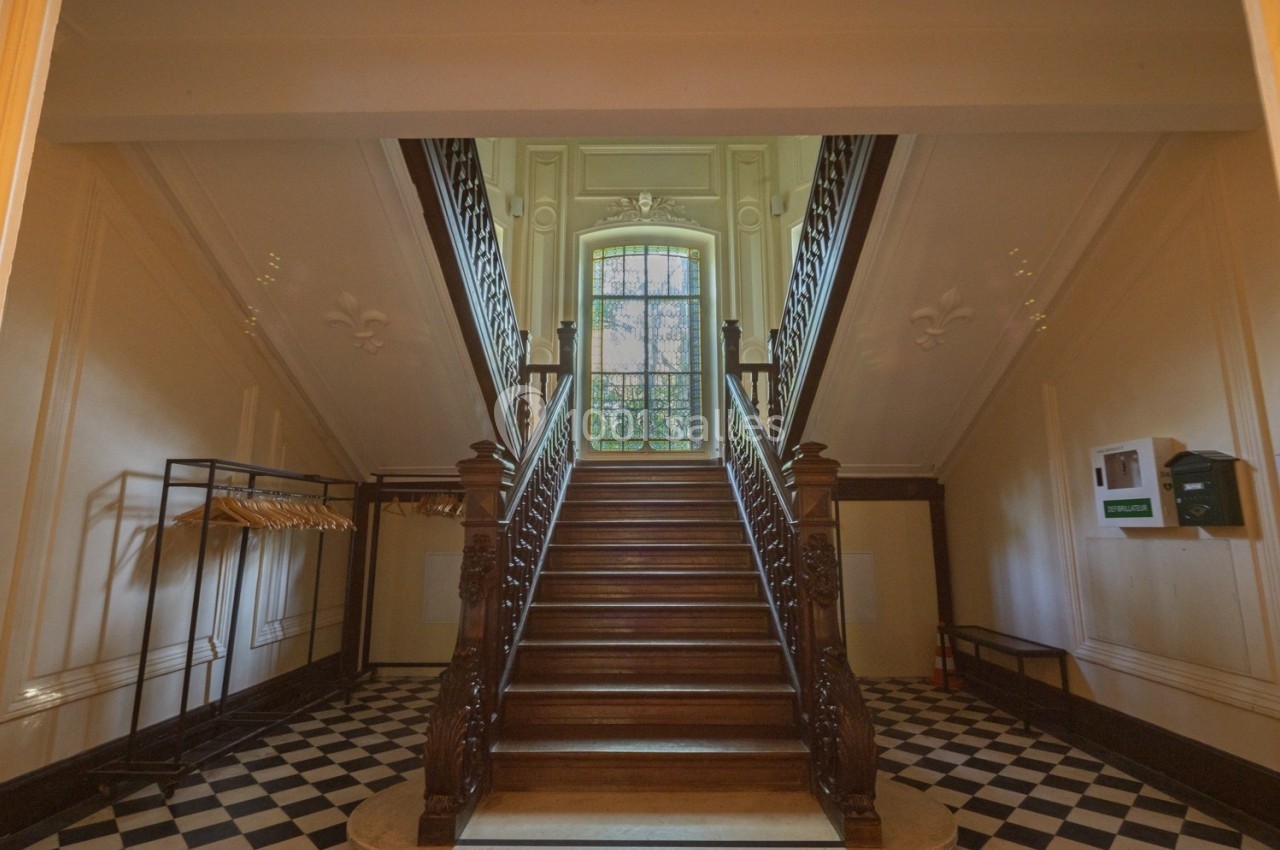 Escalier en bois massif dans un hall lumineux avec sol en damier, portant et fenêtre à vitraux en arrière-plan.