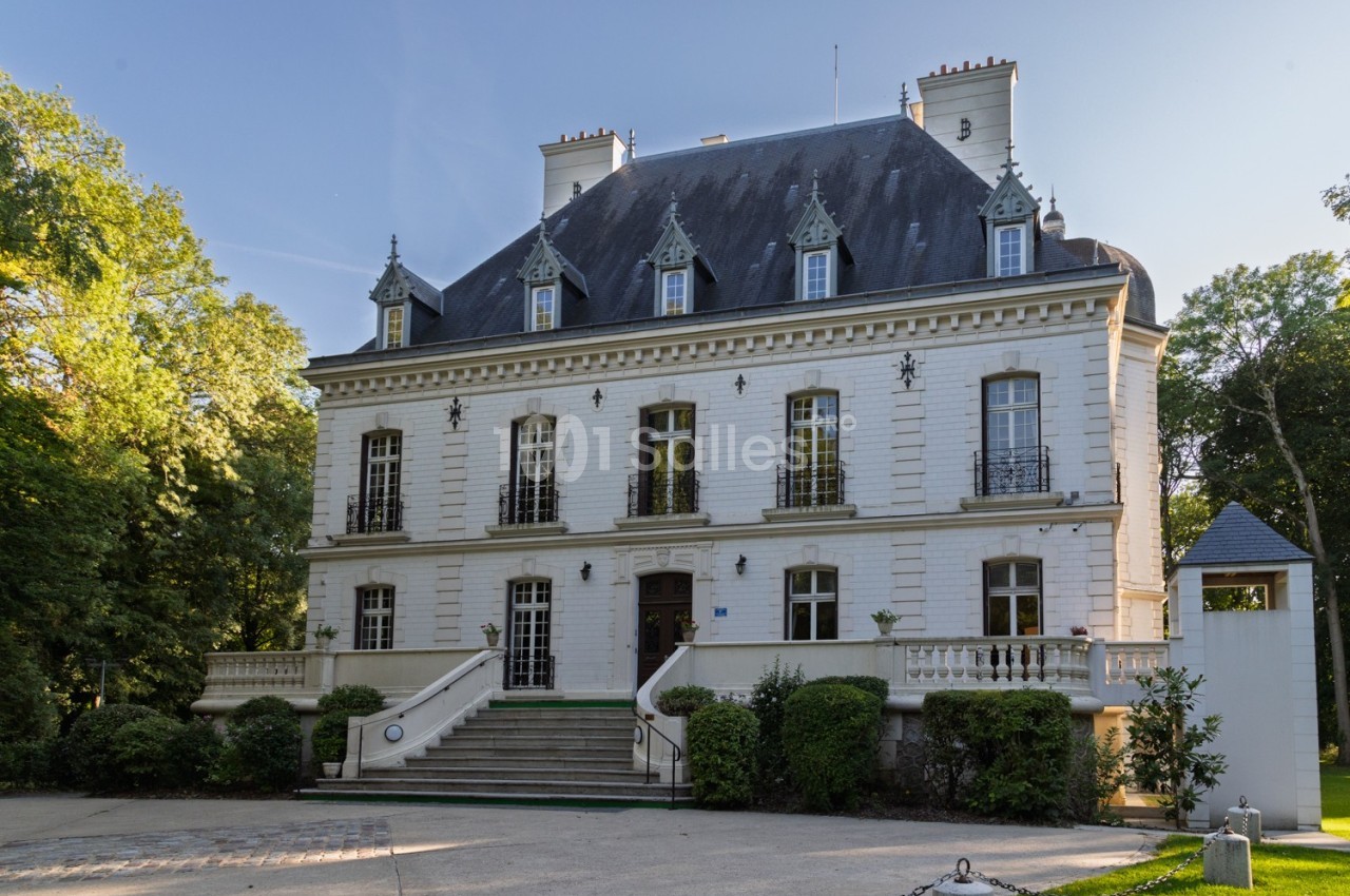 Façade d'un manoir blanc à trois étages avec toit en ardoise, entouré de verdure et d'une allée pavée.