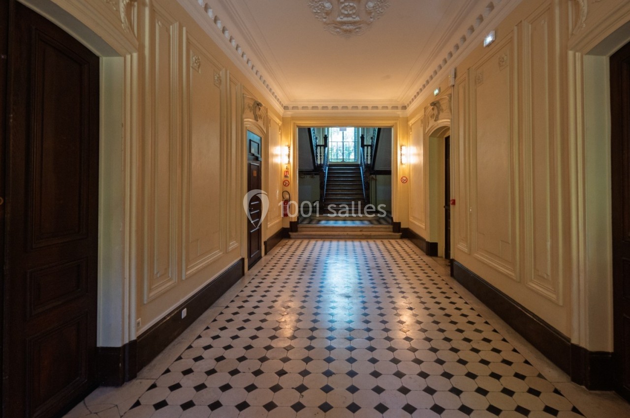 Couloir intérieur avec sol en carreaux noirs et blancs, murs ornés de moulures et escalier au fond.
