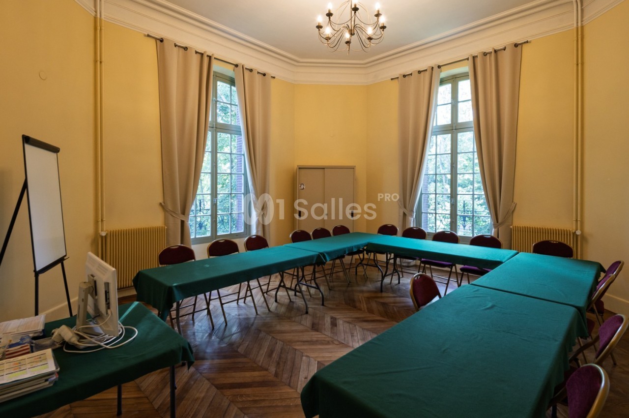 Salle de réunion lumineuse avec tables disposées en U, chaises rouges, parquet en bois et grandes fenêtres.