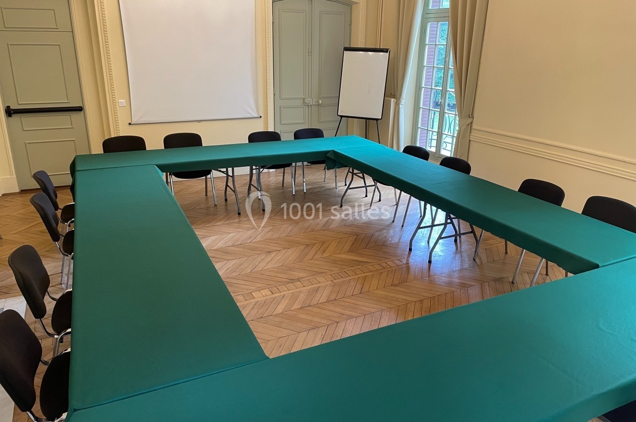 Salle de réunion avec tables disposées en U, nappes vertes, chaises noires et un tableau blanc en arrière-plan.