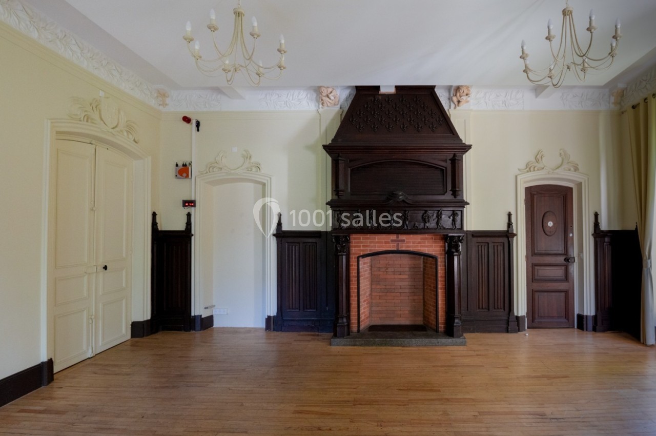 Grande pièce avec parquet, cheminée en bois sculpté, murs clairs, moulures décoratives et lustres au plafond.