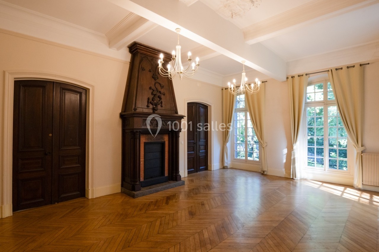 Salon lumineux avec parquet en chevrons, cheminée en bois sculpté, lustres et grandes fenêtres avec rideaux clairs.