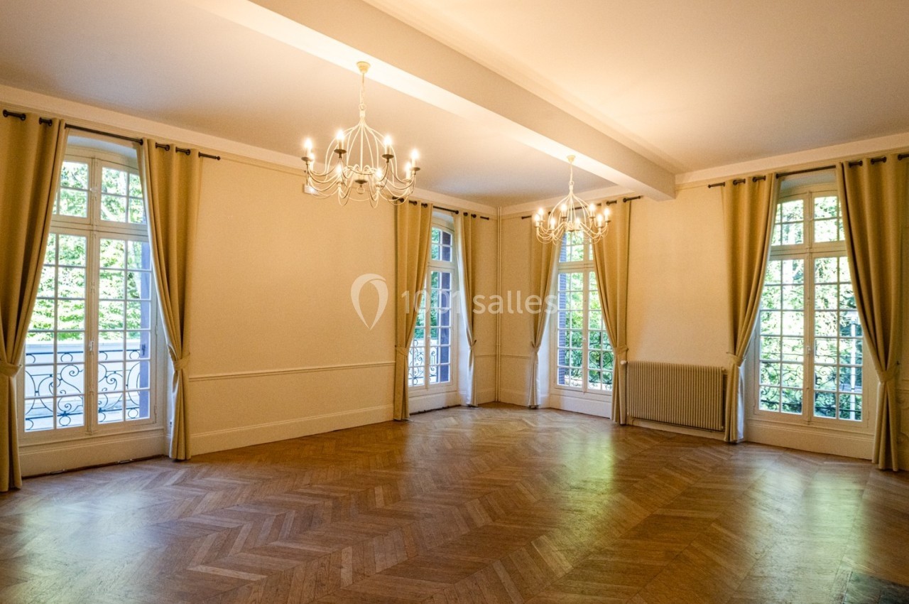 Salle lumineuse avec parquet en chevrons, grandes fenêtres avec rideaux beiges et lustres suspendus.