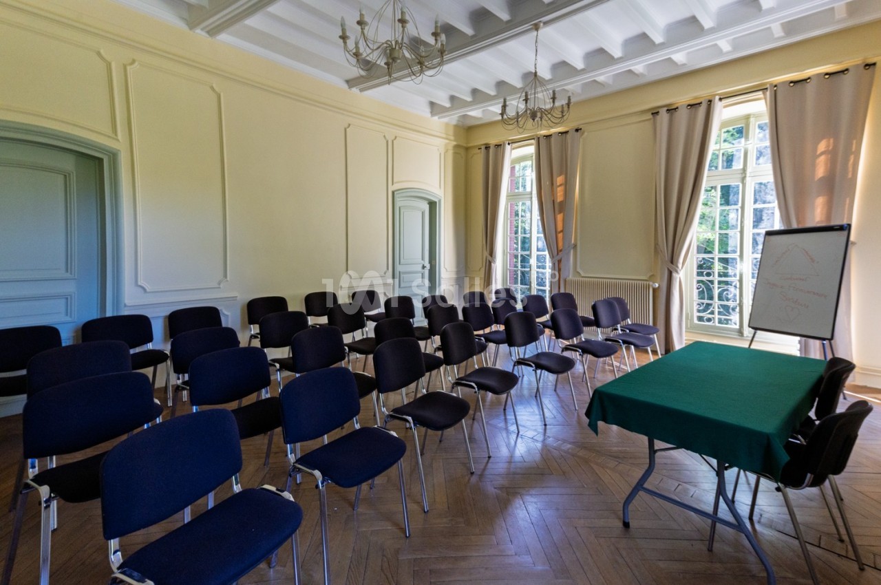 Salle lumineuse avec chaises alignées, table verte et chevalet, grandes fenêtres avec rideaux et plafond à moulures.