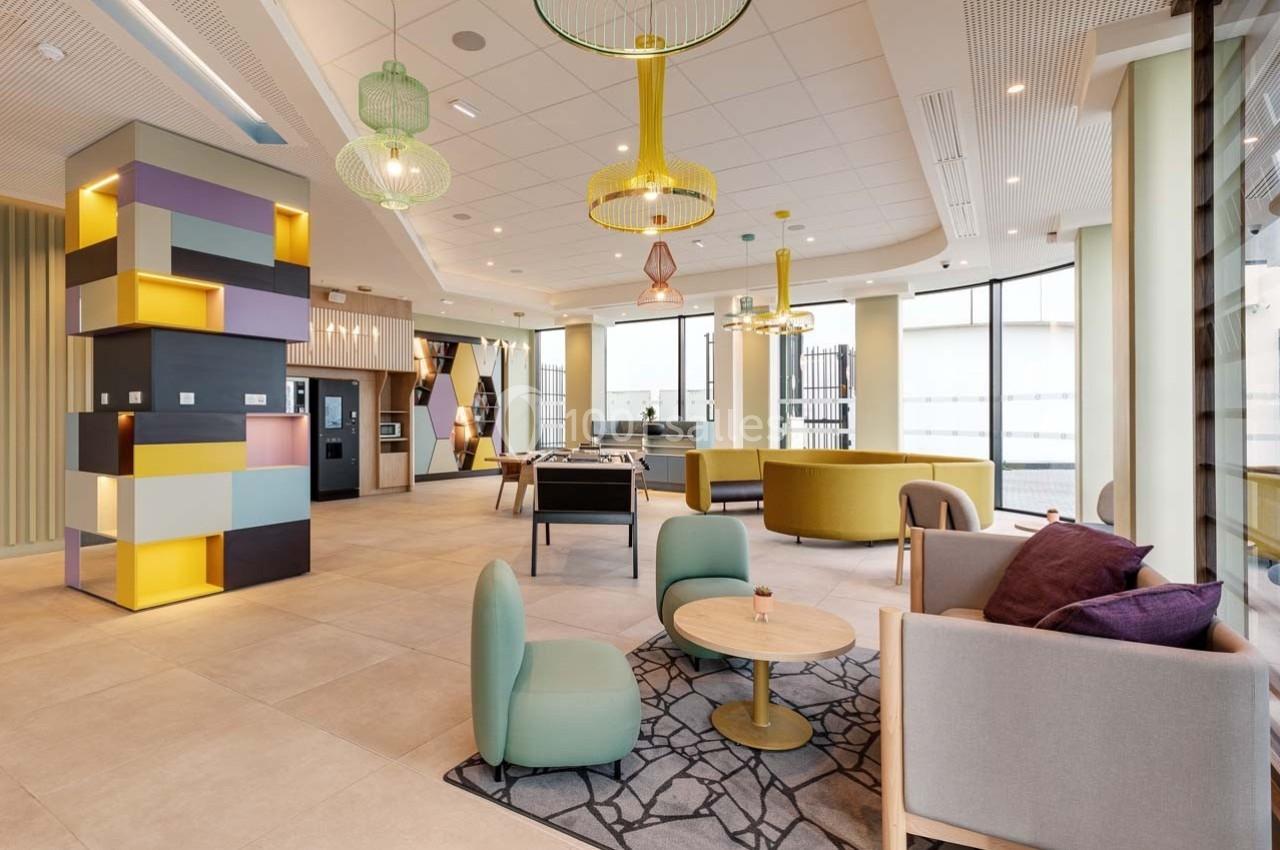 Espace intérieur lumineux avec mobilier moderne, coin salon, tables, et grandes fenêtres laissant entrer la lumière…