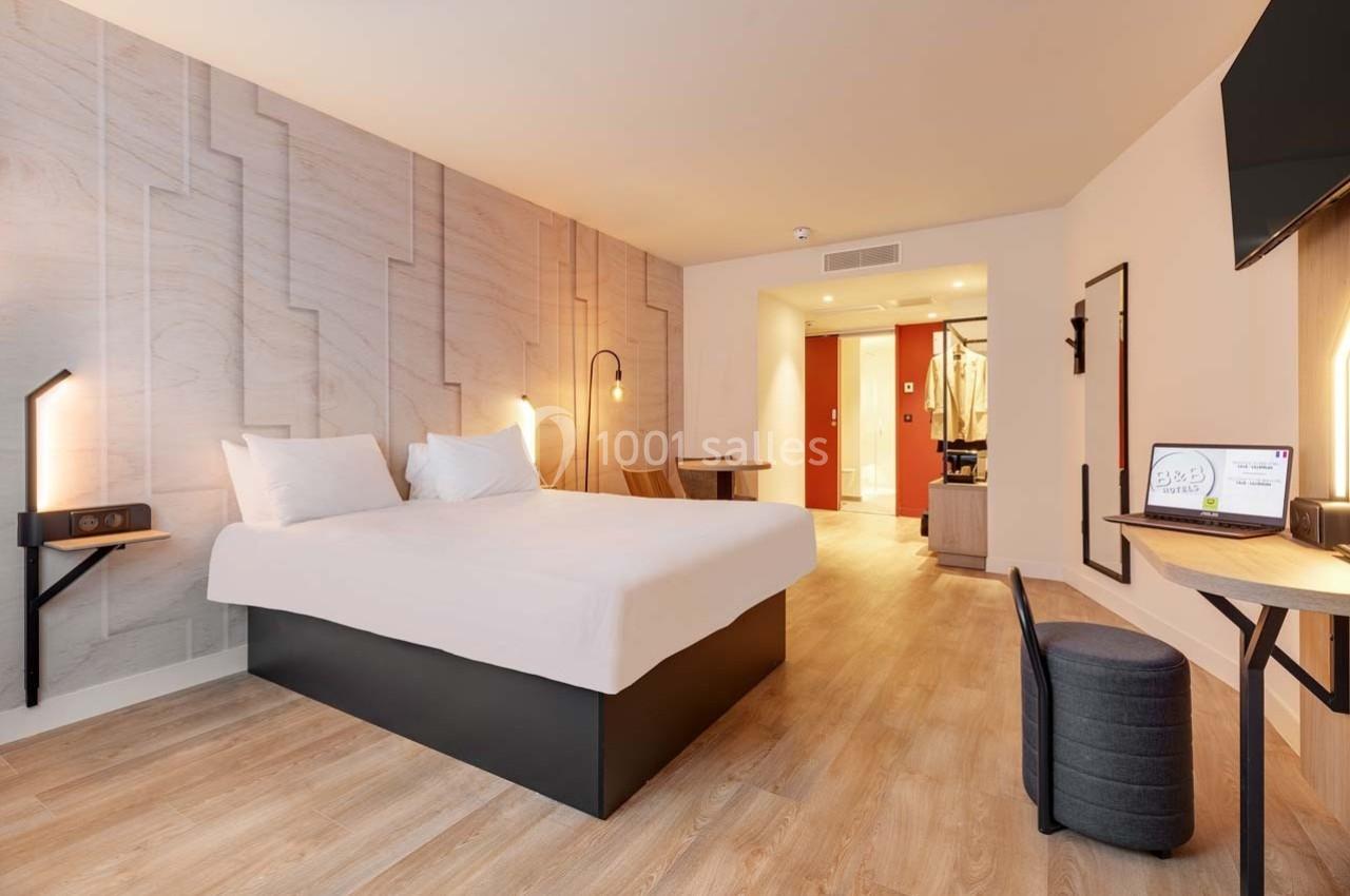 Chambre d'hôtel moderne avec lit double, bureau, télévision murale et salle de bain visible en arrière-plan.