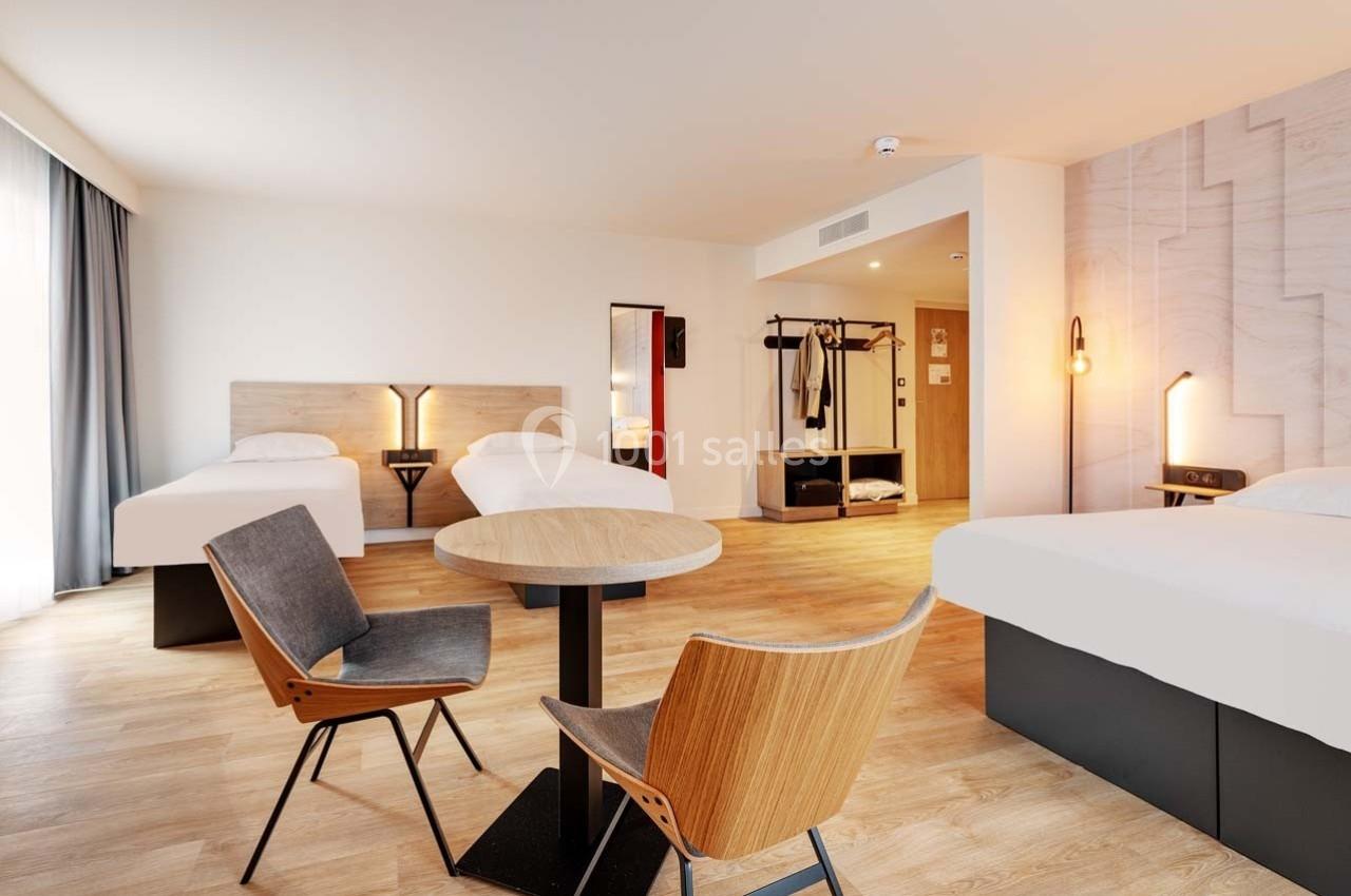 Chambre d'hôtel lumineuse avec trois lits, table ronde, chaises modernes et espace de rangement ouvert.