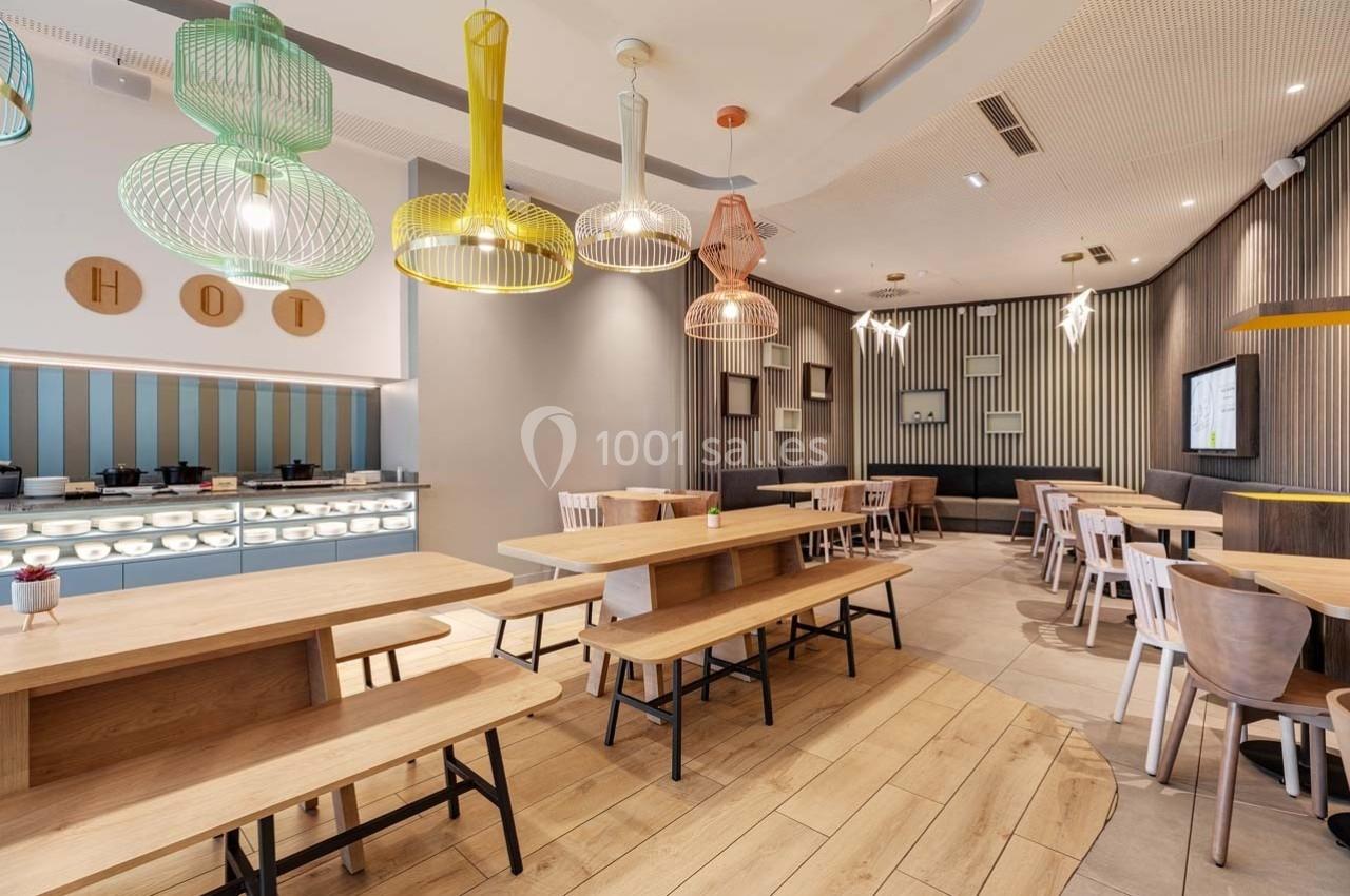 Salle de restaurant moderne avec tables en bois, chaises variées et luminaires colorés suspendus au plafond.