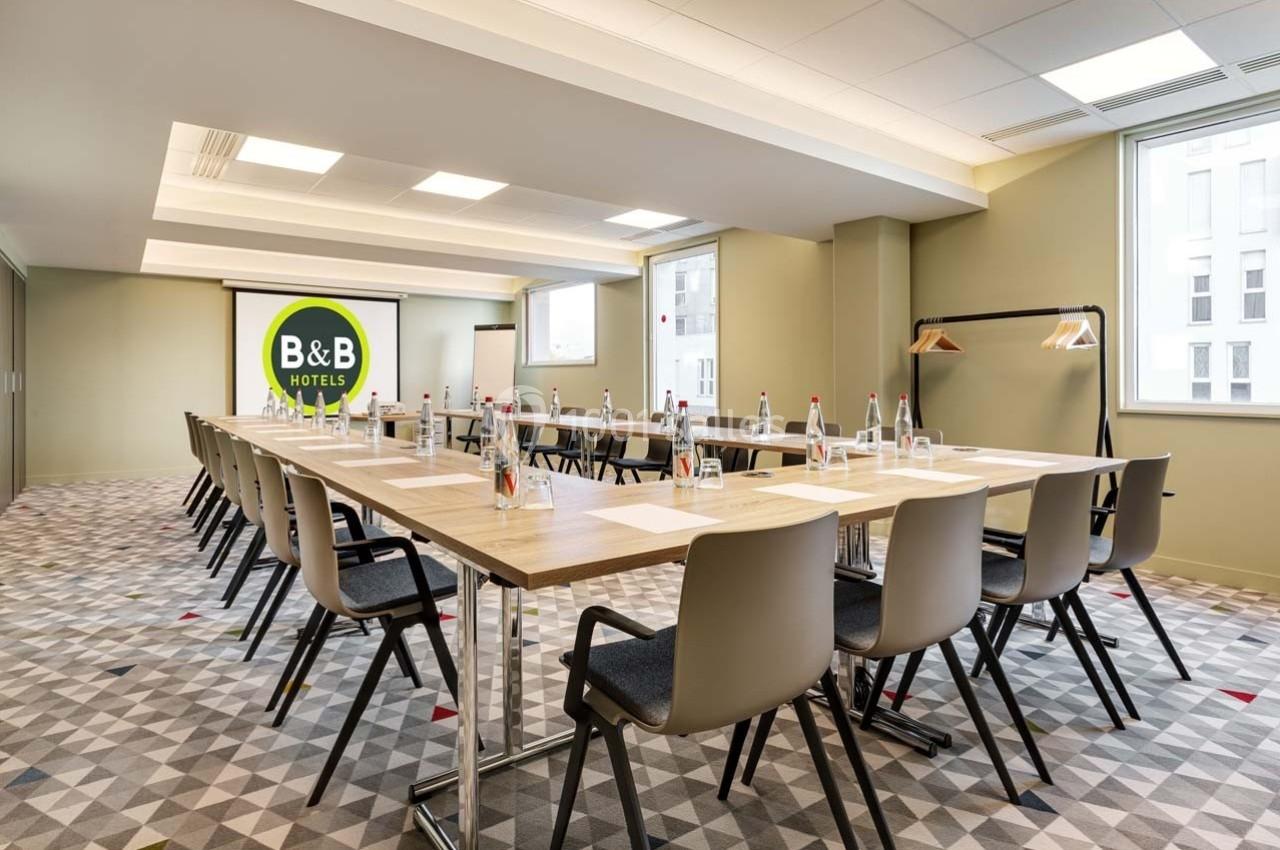 Salle de réunion lumineuse avec tables en U, chaises modernes, bouteilles d'eau et écran de présentation.