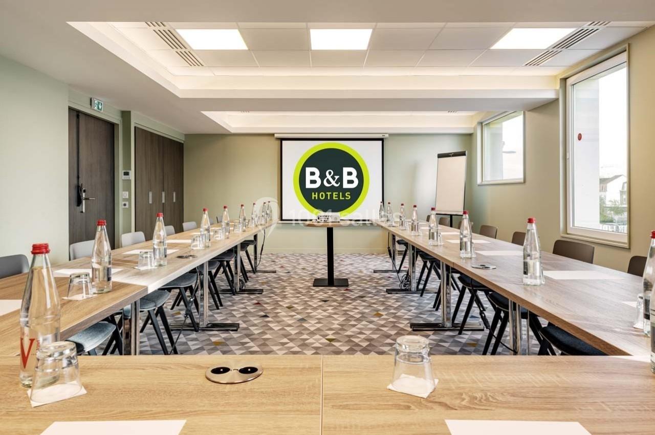 Salle de réunion lumineuse avec tables disposées en U, écran de projection et bouteilles d'eau alignées.