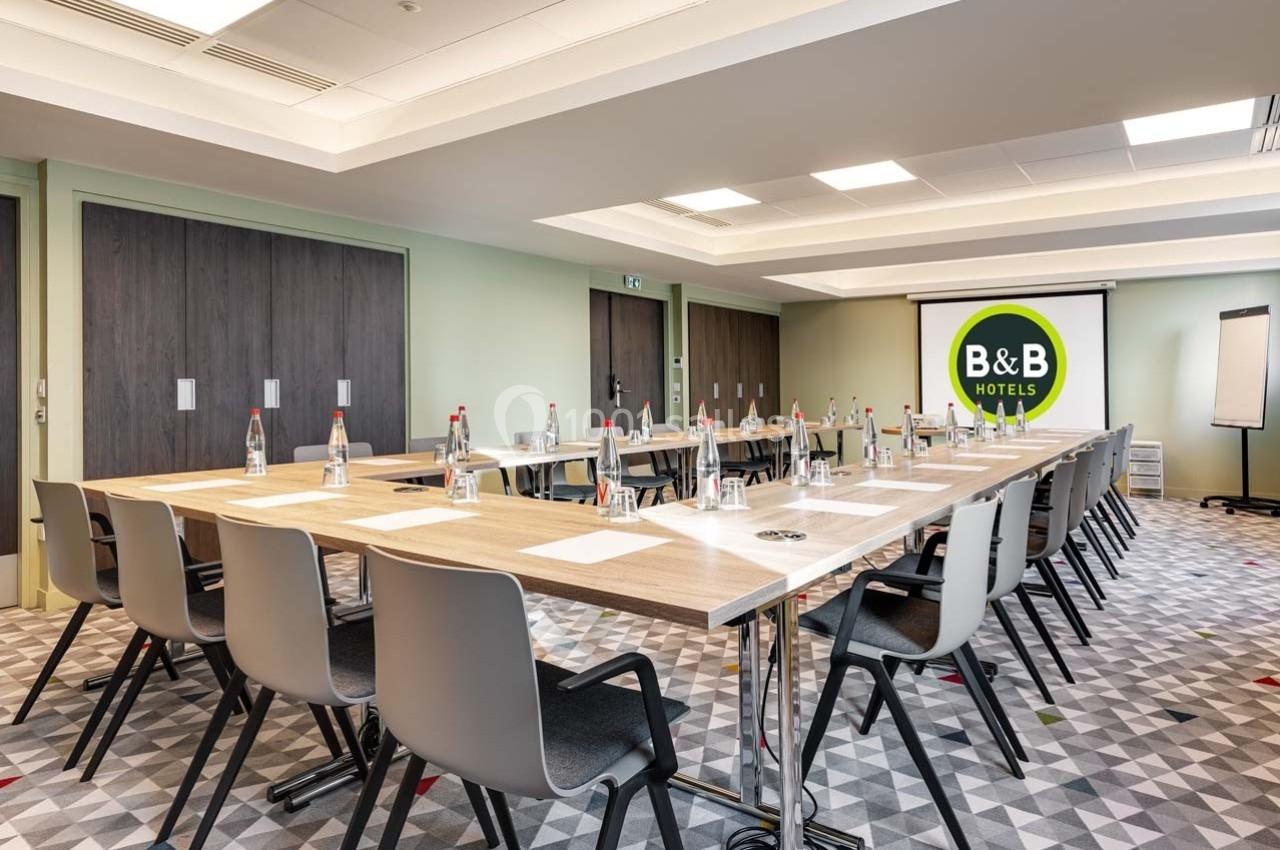 Salle de réunion lumineuse avec tables en U, chaises, bouteilles d'eau, bloc-notes et écran affichant le logo B&B Hotels.