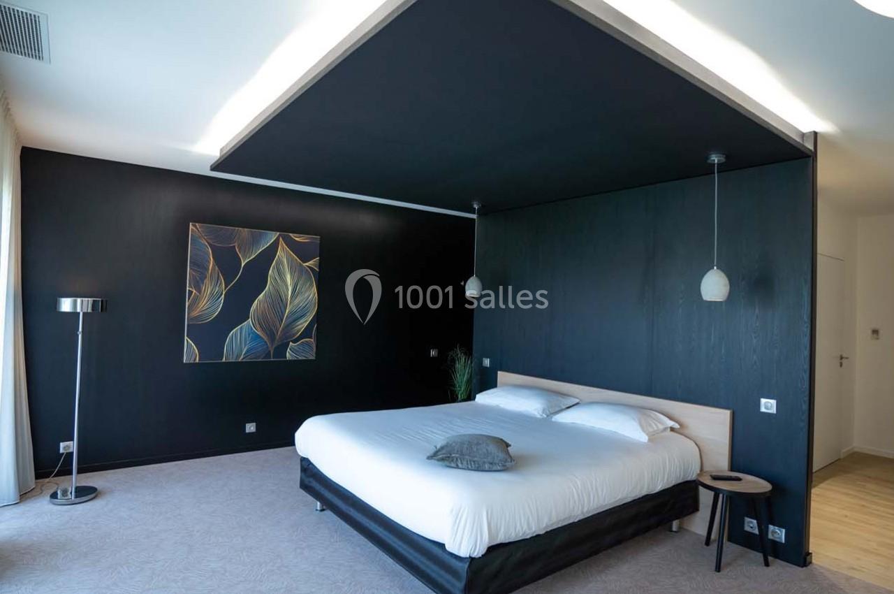 Chambre moderne avec lit double, murs sombres, tableau décoratif, lampes suspendues et grande fenêtre à gauche.