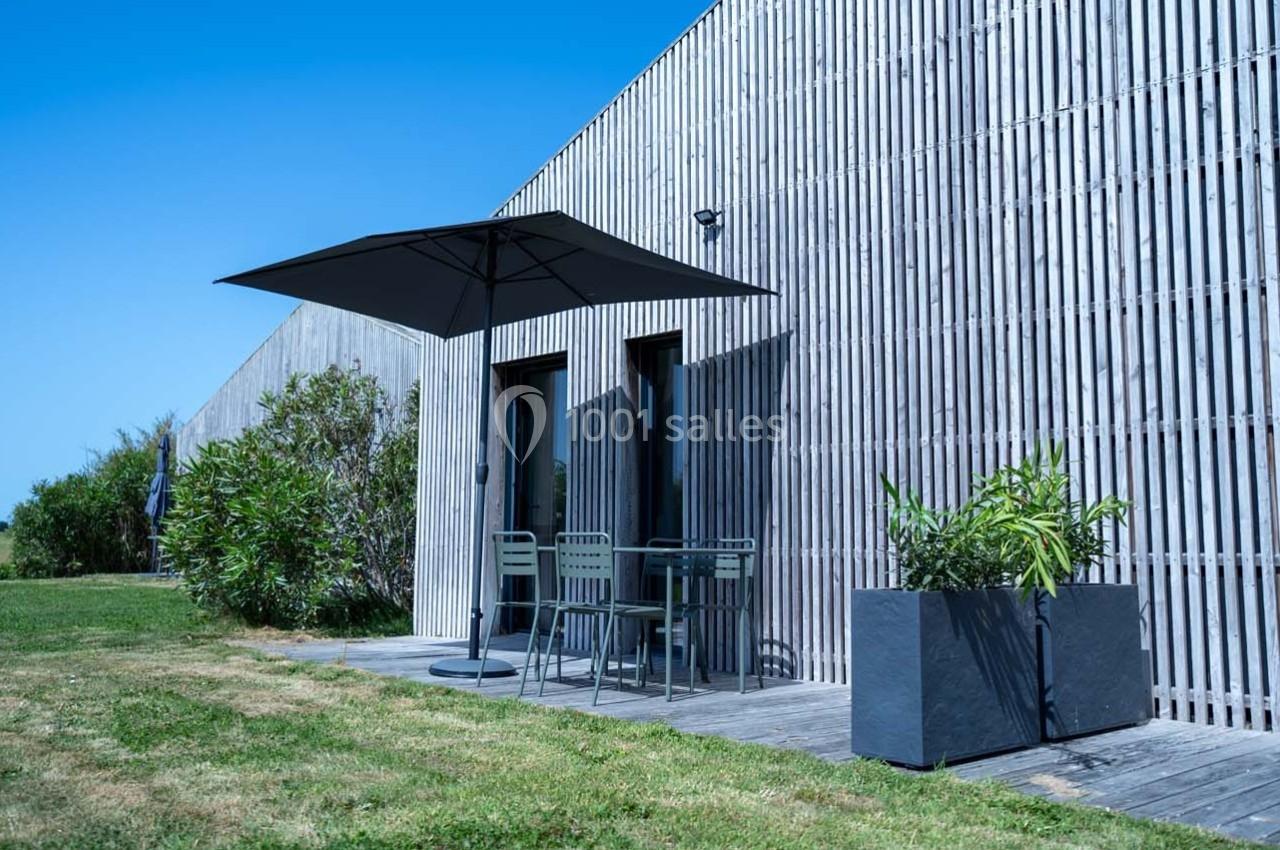 Terrasse en bois avec table, chaises et parasol, entourée de plantes, devant une façade en bois sous un ciel bleu.