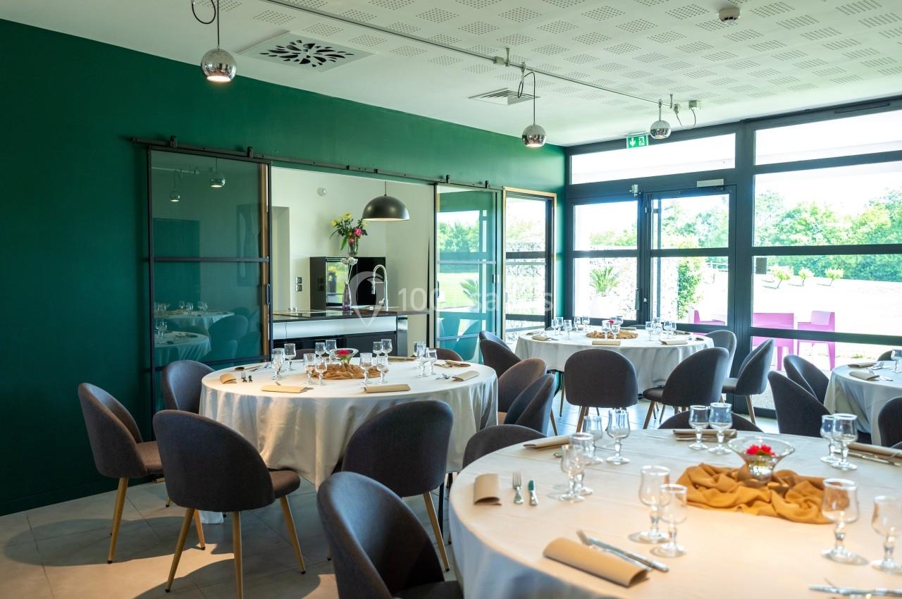 Salle lumineuse avec tables rondes dressées, chaises grises, grandes baies vitrées et murs verts.
