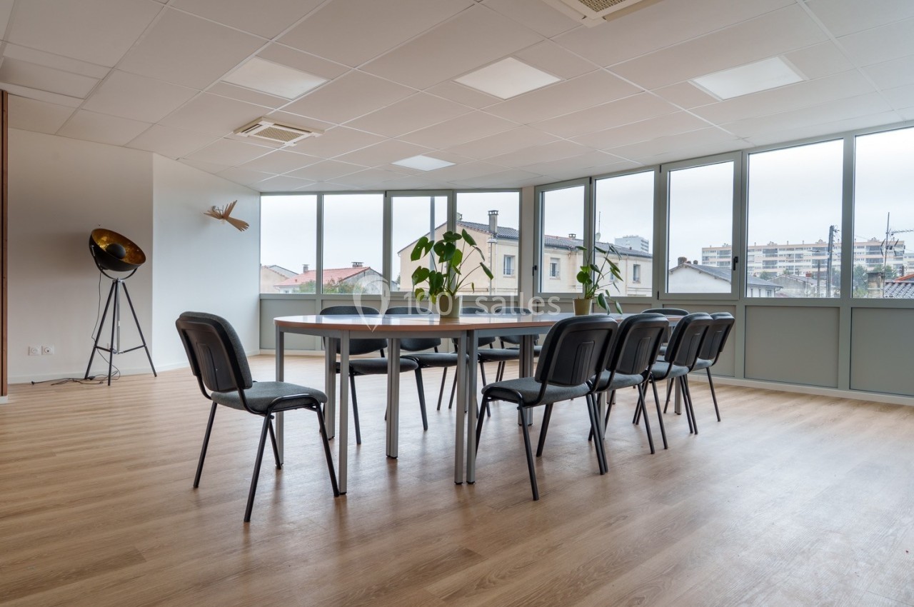 Salle de réunion lumineuse avec table rectangulaire, chaises noires, sol en bois et grandes fenêtres donnant sur des…
