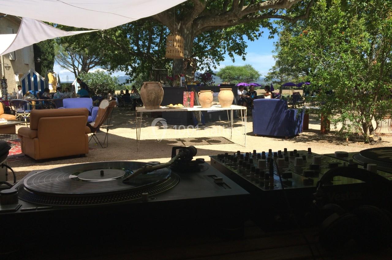 Platines de DJ en premier plan avec une terrasse ombragée, des tables et des arbres en arrière-plan.