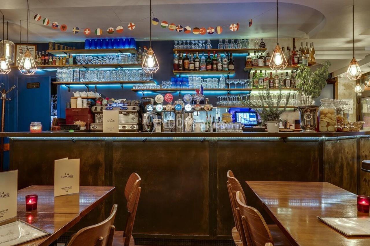 Bar avec comptoir éclairé, étagères garnies de bouteilles, tables en bois et décorations lumineuses suspendues.