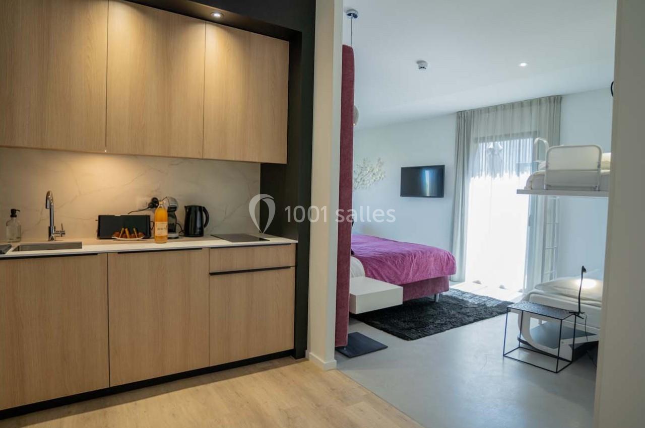 Cuisine moderne avec placards en bois, ouverte sur une chambre avec lit double, lit superposé et télévision murale.