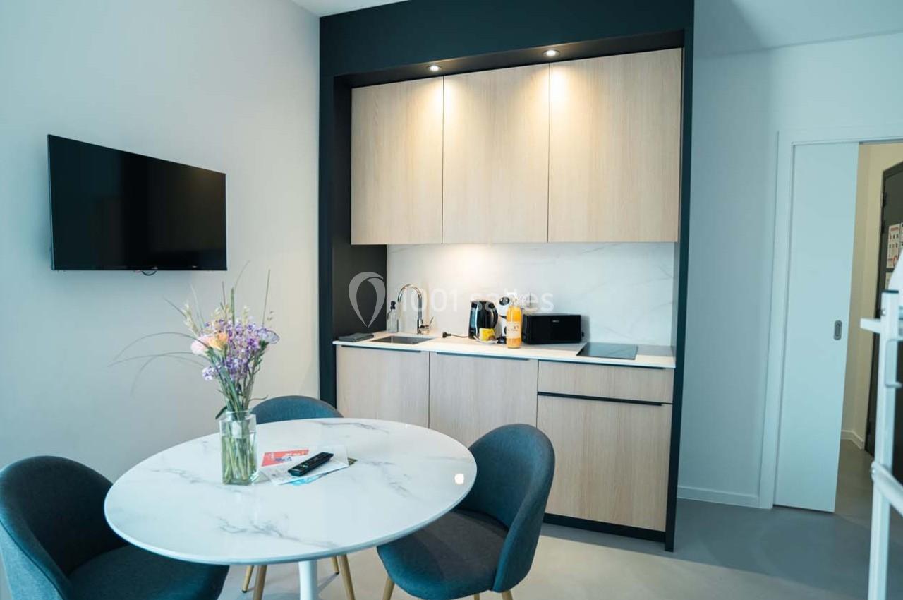 Cuisine moderne avec placards en bois clair, table ronde en marbre, chaises grises et télévision murale.