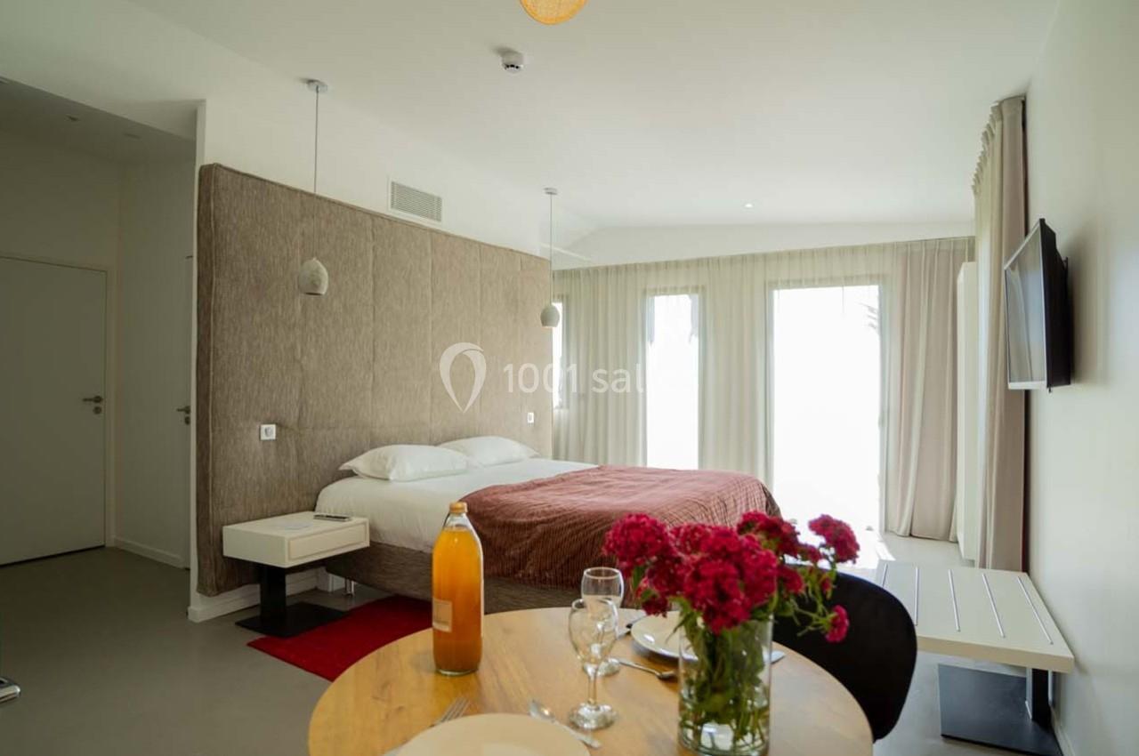 Chambre lumineuse avec lit double, table dressée, fleurs rouges, télévision murale et grandes fenêtres avec rideaux.