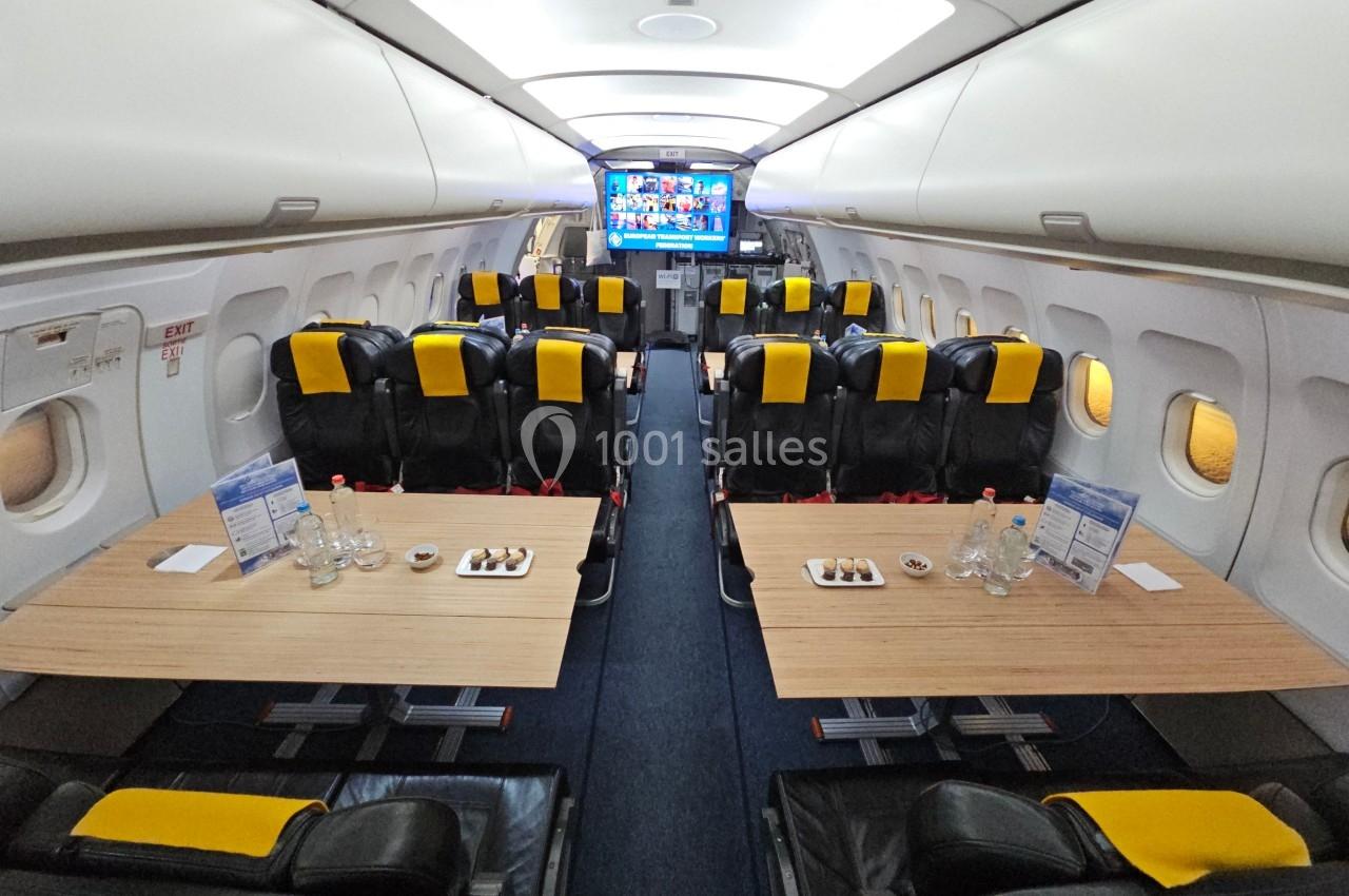 Intérieur d'un avion aménagé avec des tables en bois, des sièges noirs et jaunes, et des écrans à l'avant.