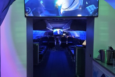 Salle d'exposition avec des stands, des écrans interactifs et des tables hautes, décorée de nuages suspendus.