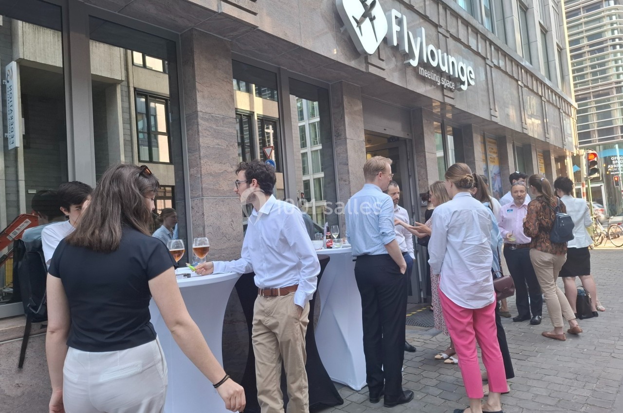 Personnes discutant debout autour de tables hautes devant un bâtiment avec le logo ’Flylounge’.