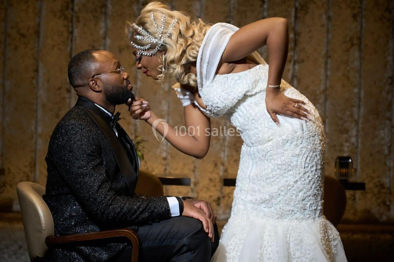 Une femme en robe blanche se penche vers un homme assis en costume noir dans un cadre intérieur élégant.