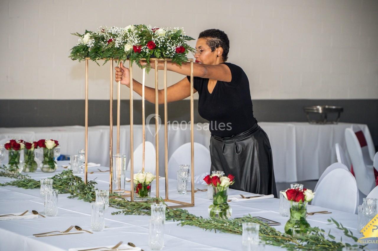 Une femme ajuste une décoration florale sur une structure métallique dans une salle de réception élégamment dressée.