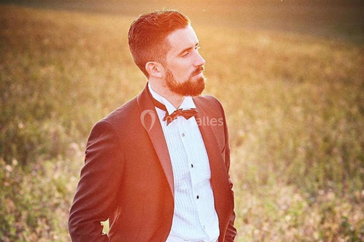 Homme en costume noir et nœud papillon, debout dans un champ au coucher du soleil.