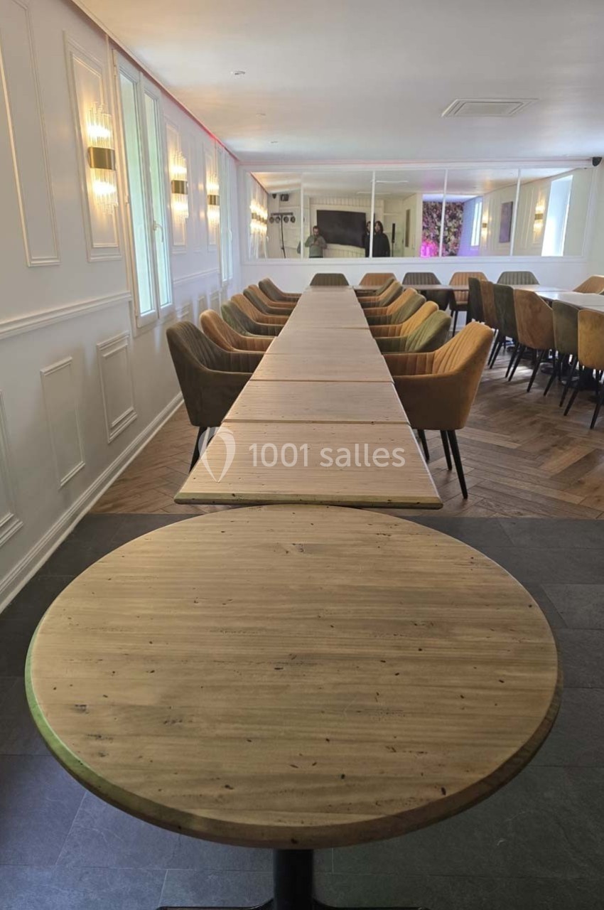 Salle lumineuse avec de longues tables en bois, entourées de chaises colorées, et un mur décoré de moulures.