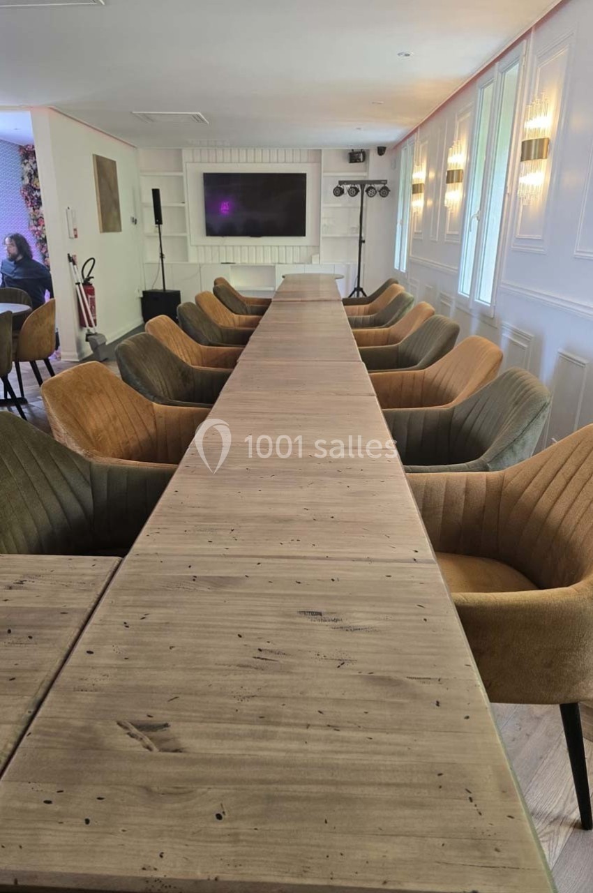 Grande table en bois entourée de fauteuils rembourrés dans une salle lumineuse avec écran et éclairages en arrière-plan.