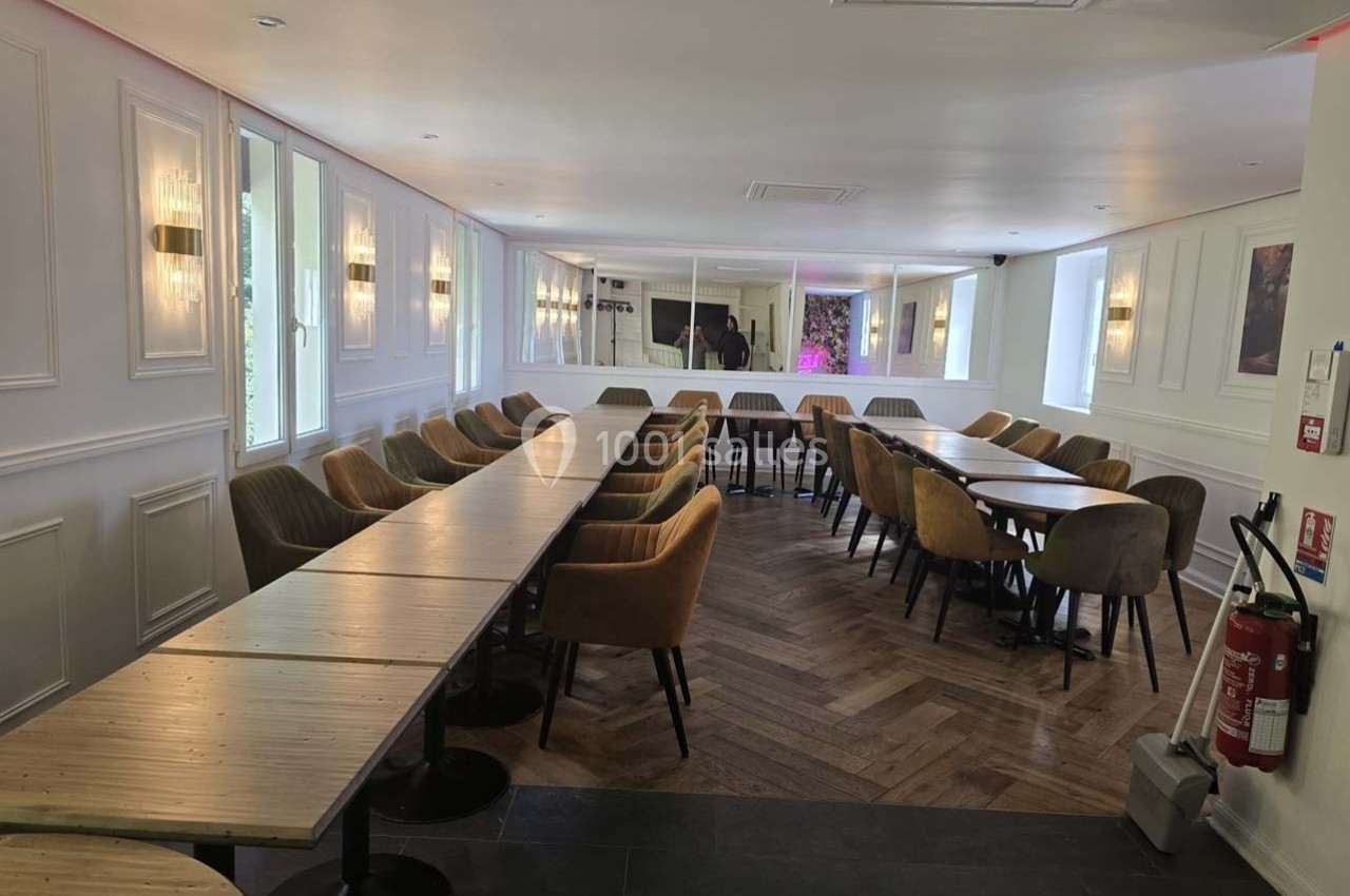 Salle lumineuse avec de longues tables en bois, chaises rembourrées, parquet en chevrons et murs blancs ornés de moulures.