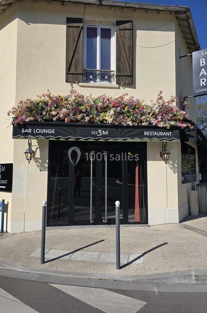Façade d'un bar-restaurant avec terrasse fleurie, enseigne ’Les 3M’, situé dans un bâtiment à volets en bois.