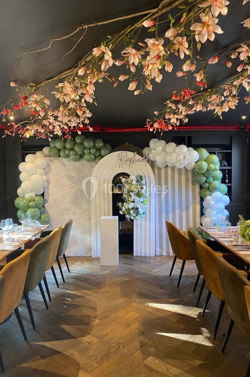 Décoration de salle avec arche florale, ballons verts et blancs, et tables dressées sous des branches fleuries.
