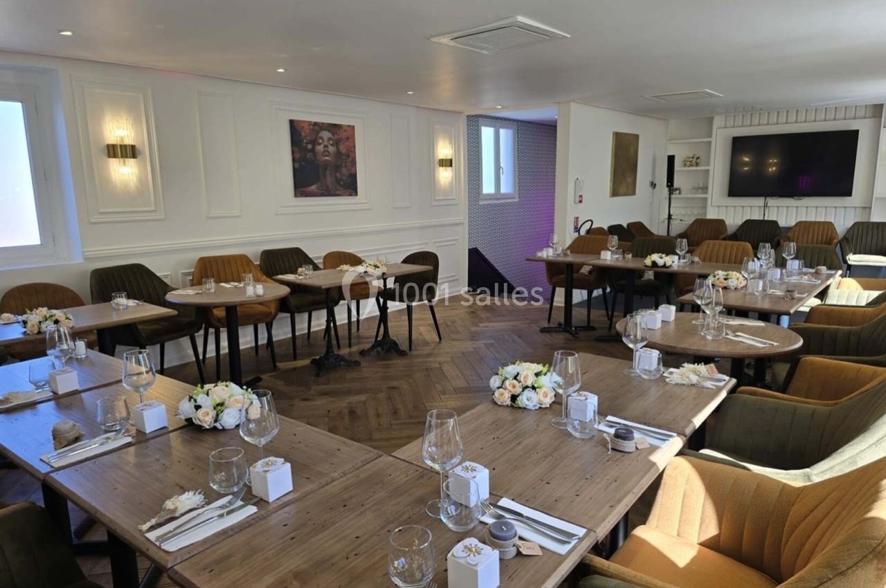 Salle lumineuse aménagée avec des tables en bois, chaises confortables et décorations florales pour un événement.